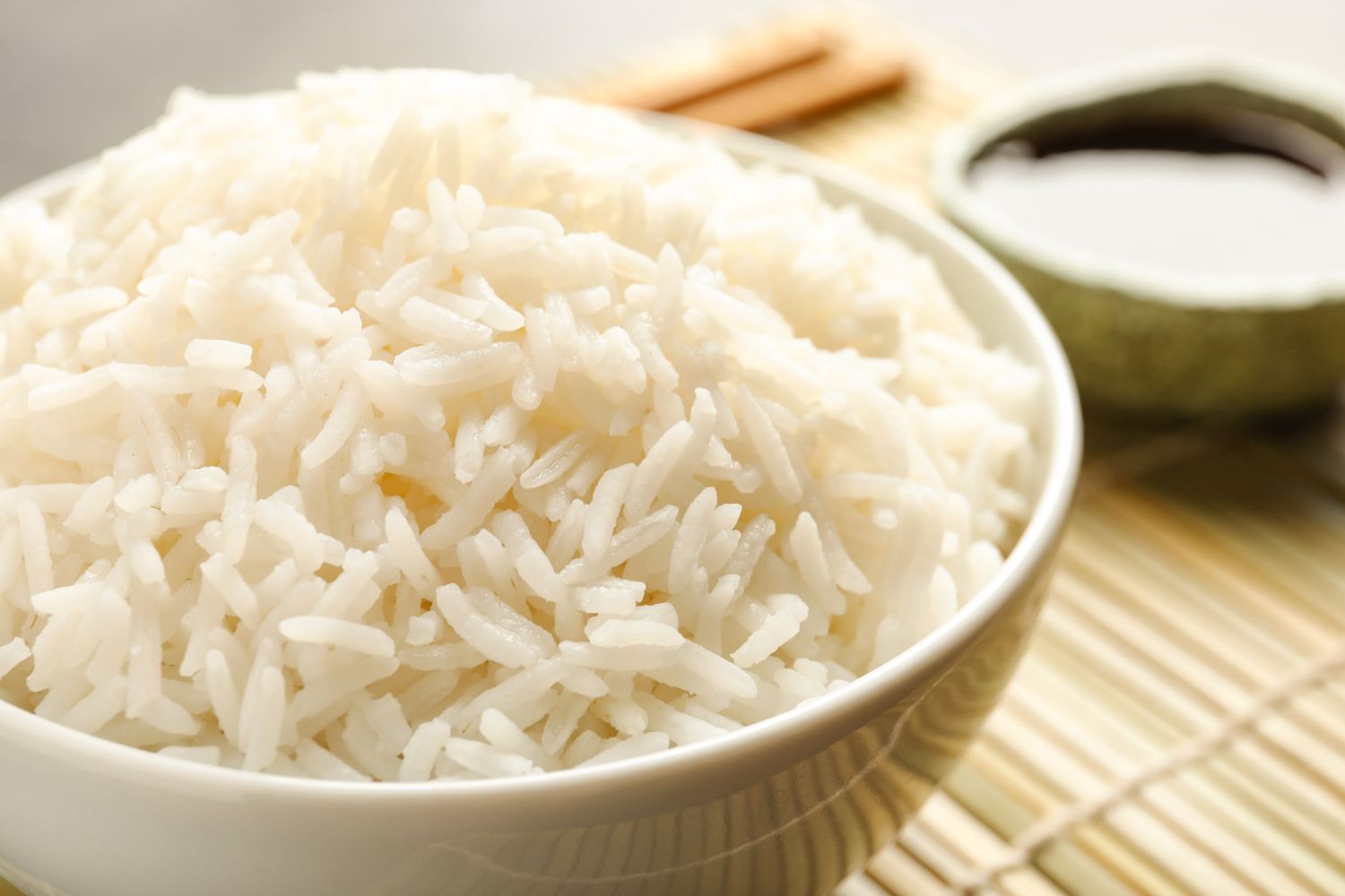 Bowl of cooked rice on a bamboo mat with a small container of sauce in the background