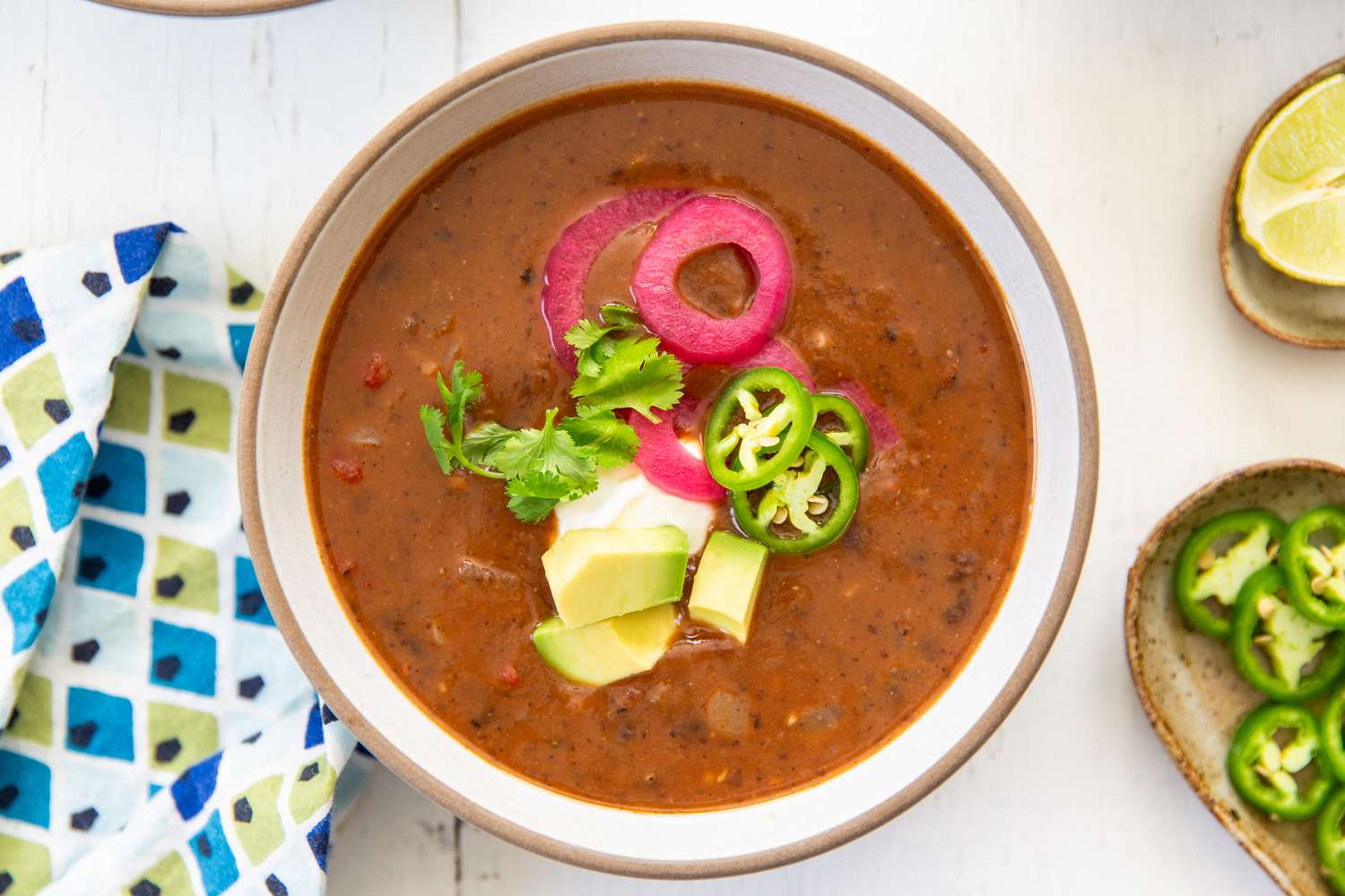 Overhead shot of a bowl of 15-minute black bean soup, topped with avocado cubes, jalapeño slices, pickled onion and and cilantro