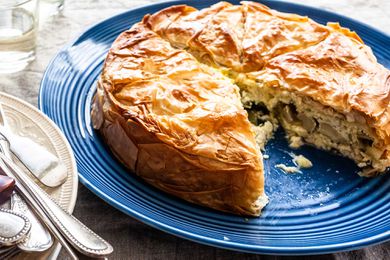 Side view of a flaky spring pie on a large blue platter. One slice of the pie is missing and the cheesy artichoke filling is visible on the plate. The lower left has a pile of forks on a white plate. A grey linen tablecloth is on the table and a glass is in the upper left corner.