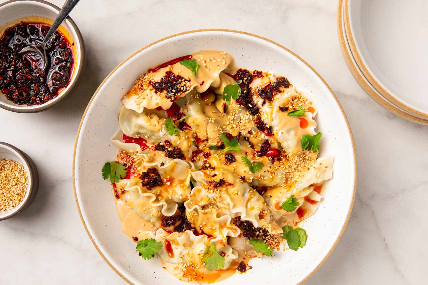 A plate of dumplings topped with peanut-chili sauce garnished with herbs and sesame seeds accompanied by a small bowl of chili oil