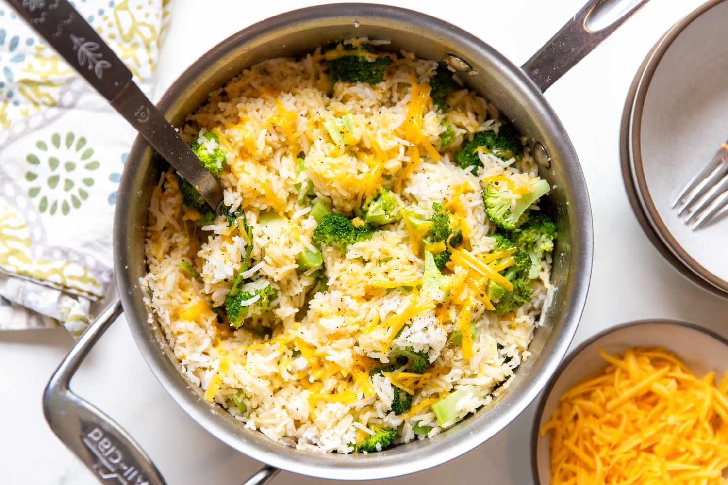 A pot of broccoli and cheese mixed with rice served alongside a small bowl of shredded cheese