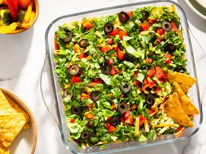 Layered nacho dip in a glass dish with toppings like lettuce tomatoes and olives served with tortilla chips