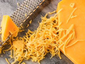 Overhead view of a large block of cheddar cheese, a shredder and pile of shredded cheese on a marble countertop