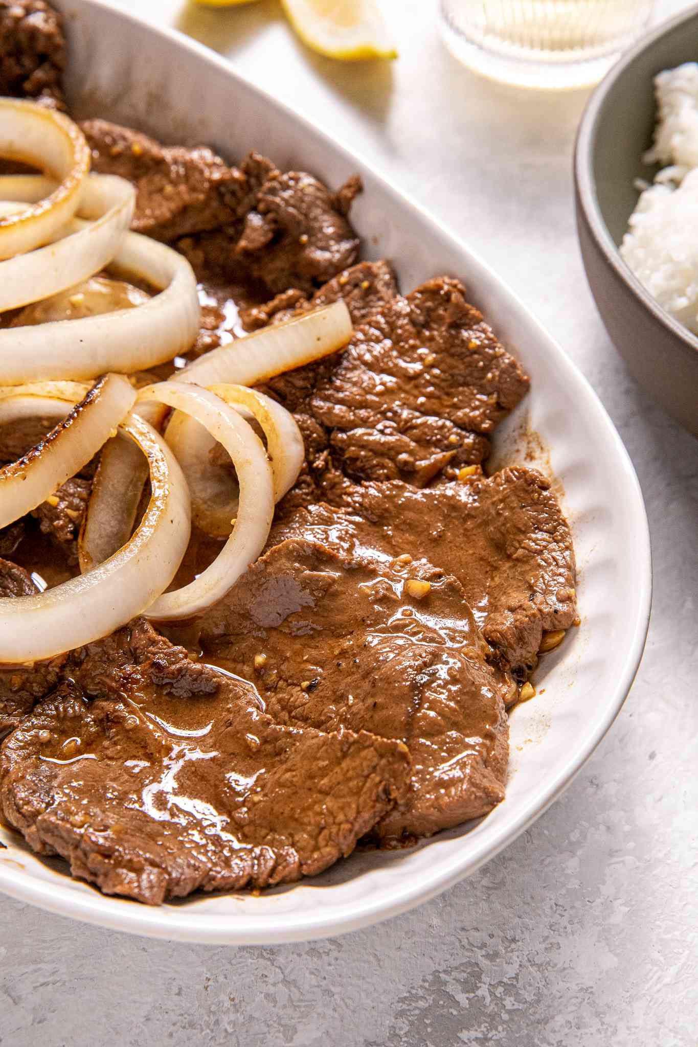 Plate of Bistek Topped With Onions Next to a Bowl of Rice