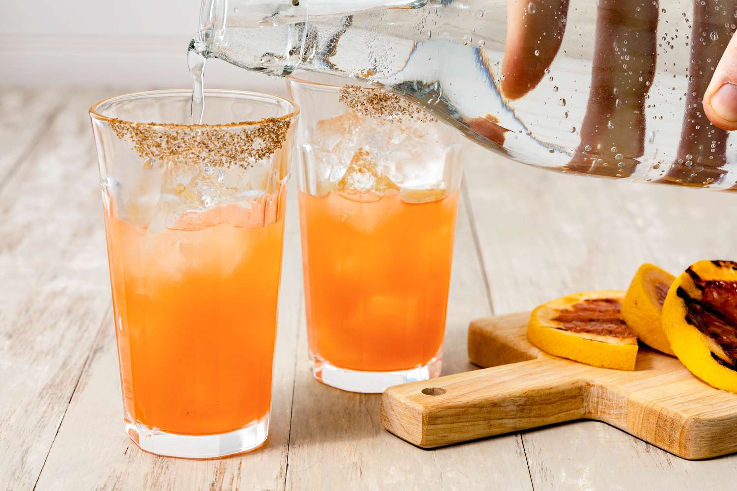 Salt Rimmed Glass of Grilled Grapefruit Paloma Topped With Club Soda Next to Another Glass and a Cutting Board With Sliced Grapefruit