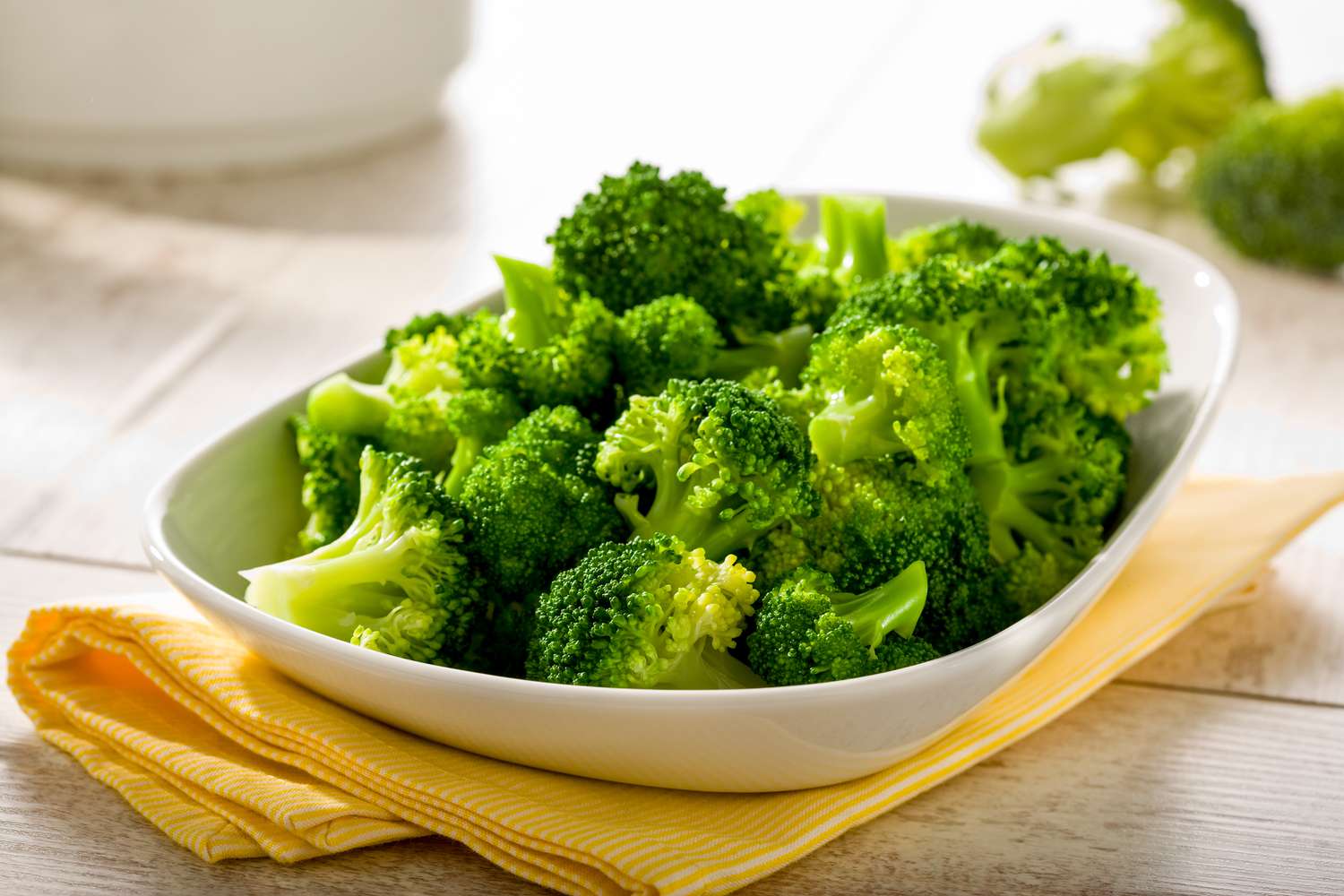 Angled view of a white bowl of steamed broccoli on top of a yellow cloth napkin and wooden tabletop