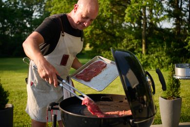 Person grilling meat using tongs on a charcoal grill in a garden setting on a sunny day, wearing an apron and holding a tray