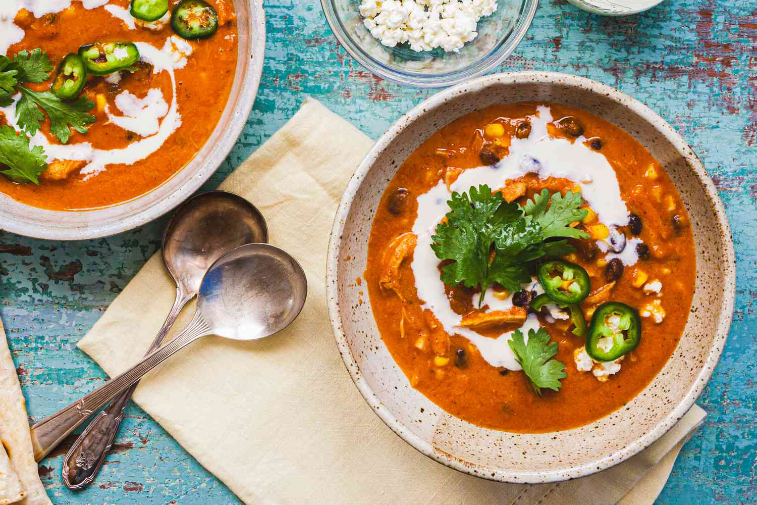 Bowls of Creamy Chicken Enchilada Soup at a Table Setting with Spoons on a Table Napkin, Tortillas in the Corner, and a Small Bowl of Cotija