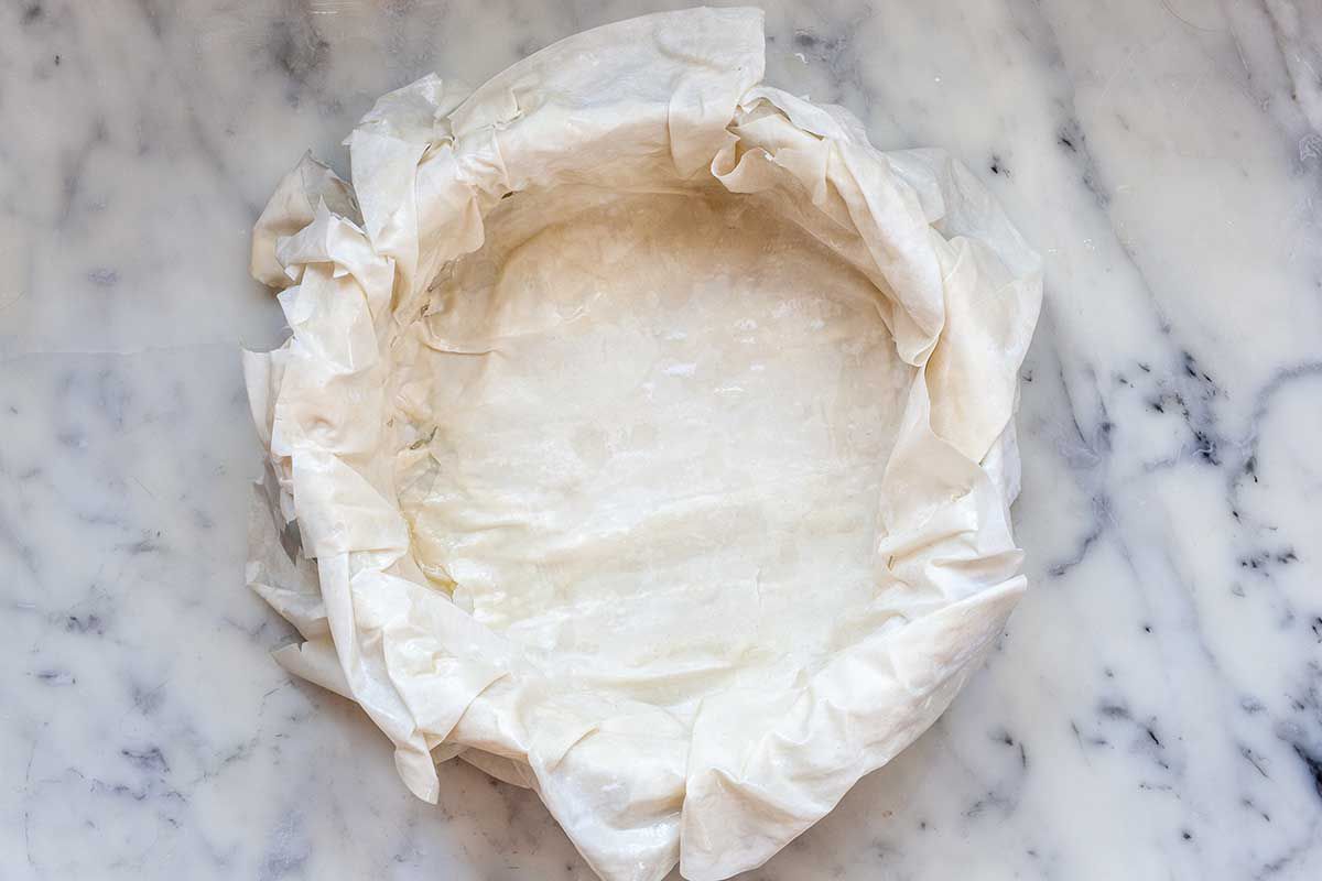 A cake pan is on a marble countertop. The phyllo is draped over the sides of the pan and covering the bottom..
