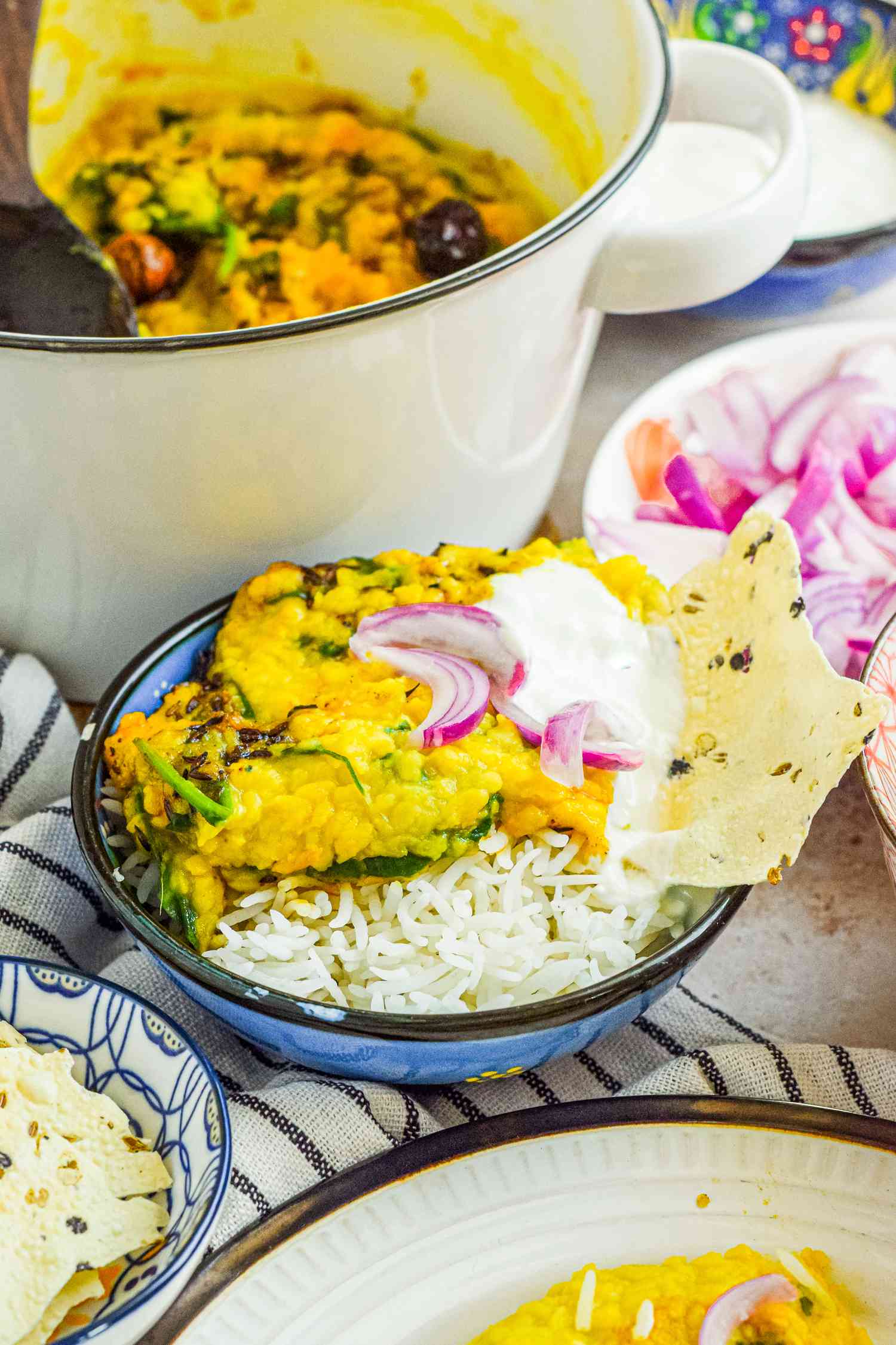 Bowl of Daal Palak over Rice Topped with Onions and Yogurt, and Served with a Piece of Papad, and in the Surroundings, More Daal Palak in a Dutch Oven, a Bowl with More Papad, a Bowl of Pickled Onions, and a Plate with Another Serving of Daal Palak