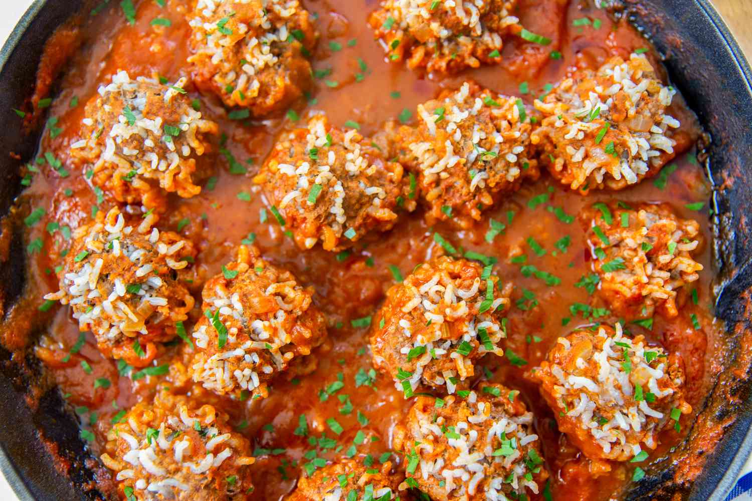 Overhead closeup view of a cast iron skillet of finished meatballs with expanded rice on the surface and sauce on a wooden cutting board