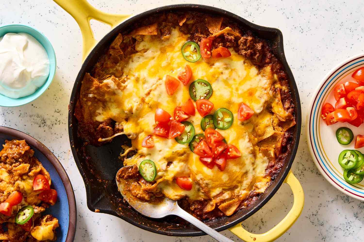 A beef enchilada skillet topped with melted cheese, chopped tomatoes, and sliced jalapenos, served with sour cream and additional toppings in dishes