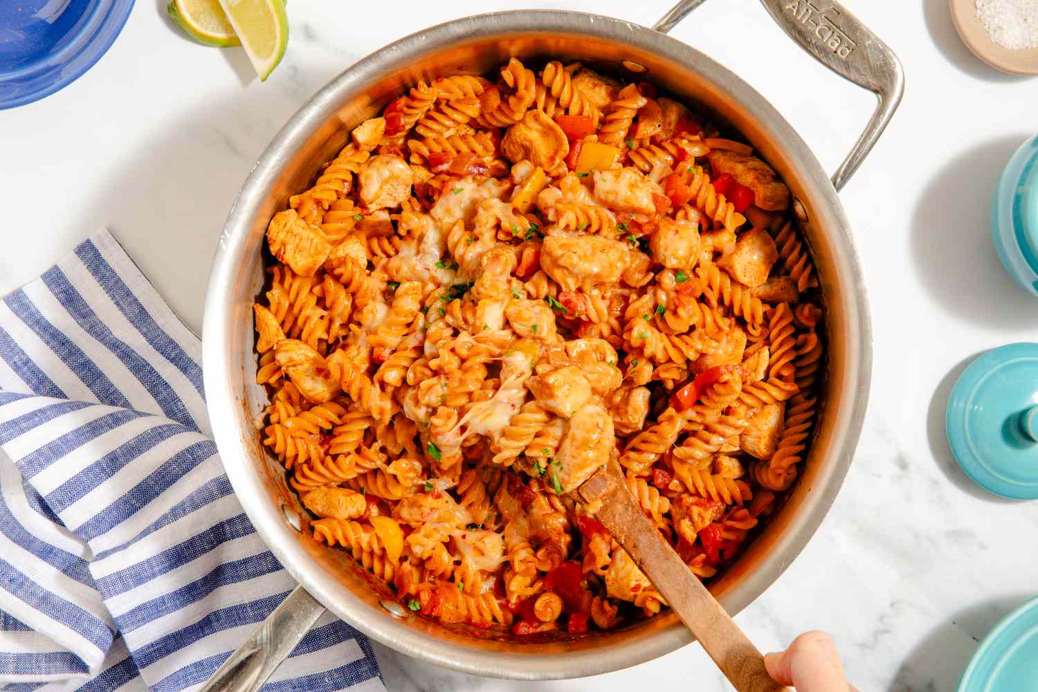 One-Pan Chicken Fajita Pasta in a pot with a hand scooping some with a wooden spoon, limes, napkins, salt on the side
