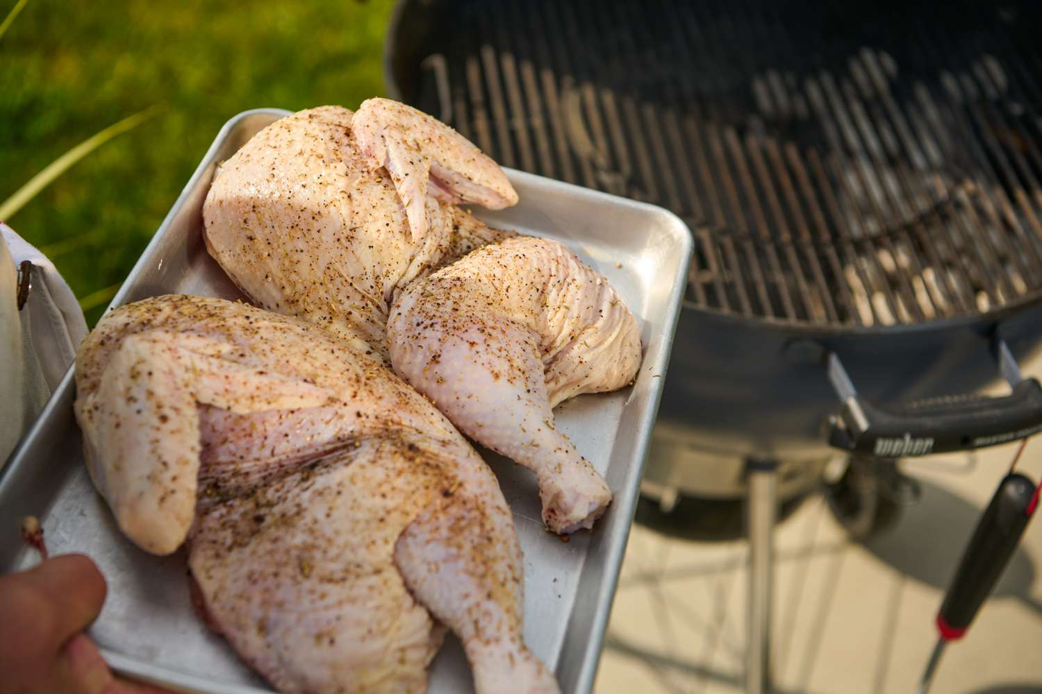 Seasoned raw chicken halves on a tray near a charcoal grill for Grilled Half Chicken recipe
