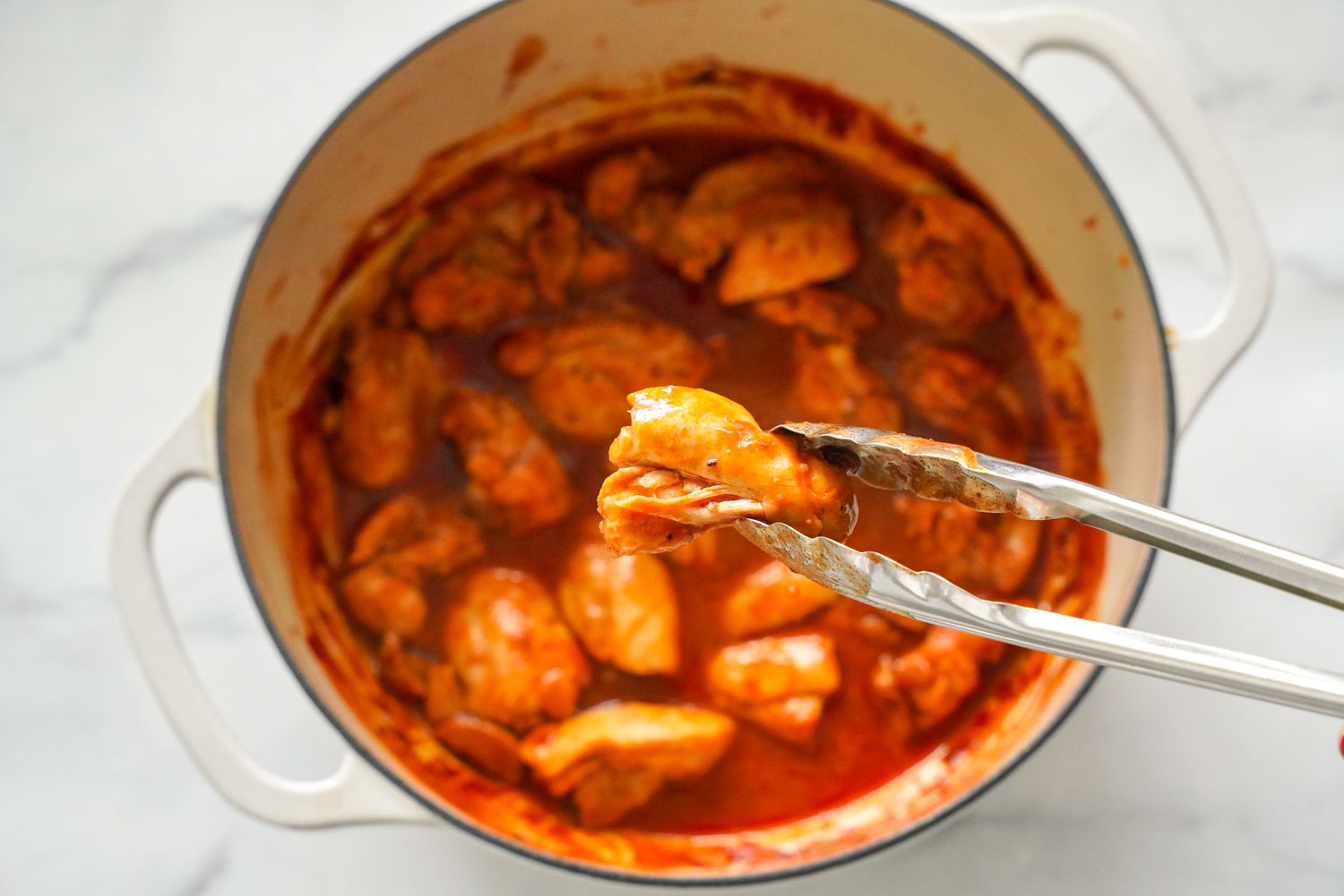 Piece of Chicken Thigh Lifted Using a Pair of Tongs From Dutch Oven With Other Cooked Pieces for Stove-top Pulled Chicken Recipe
