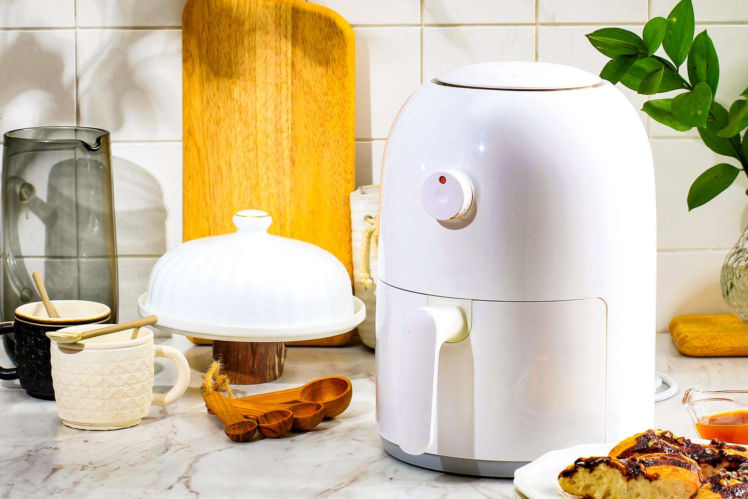 An air fryer on a kitchen counter surrounded by other kitchen tools like a set of olive wood measuring spoons, a cutting board, and two coffee mugs