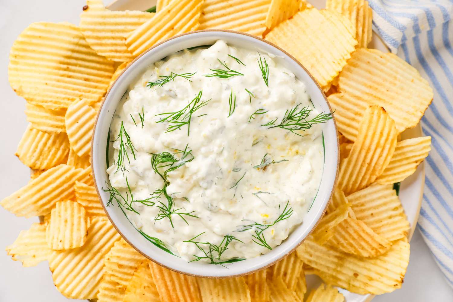 A bowl of creamy pickle dip garnished with dill, surrounded by ridged potato chips