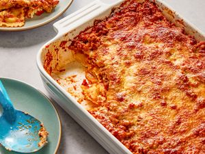 Angled view of a white baking dish of Million Dollar Ravioli Casserole with a serving removed and on a small plate next to a serving spoon on a small plate