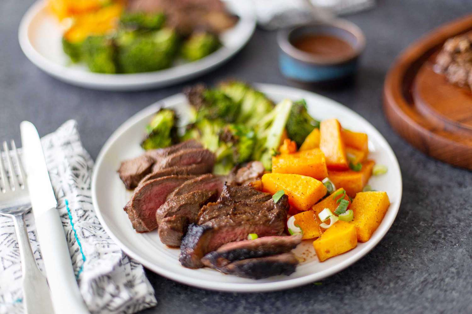 Side view of two plates with sliced easy broiled steak, chopped broccoli and squash. Silverware and a linen are next to each plate.