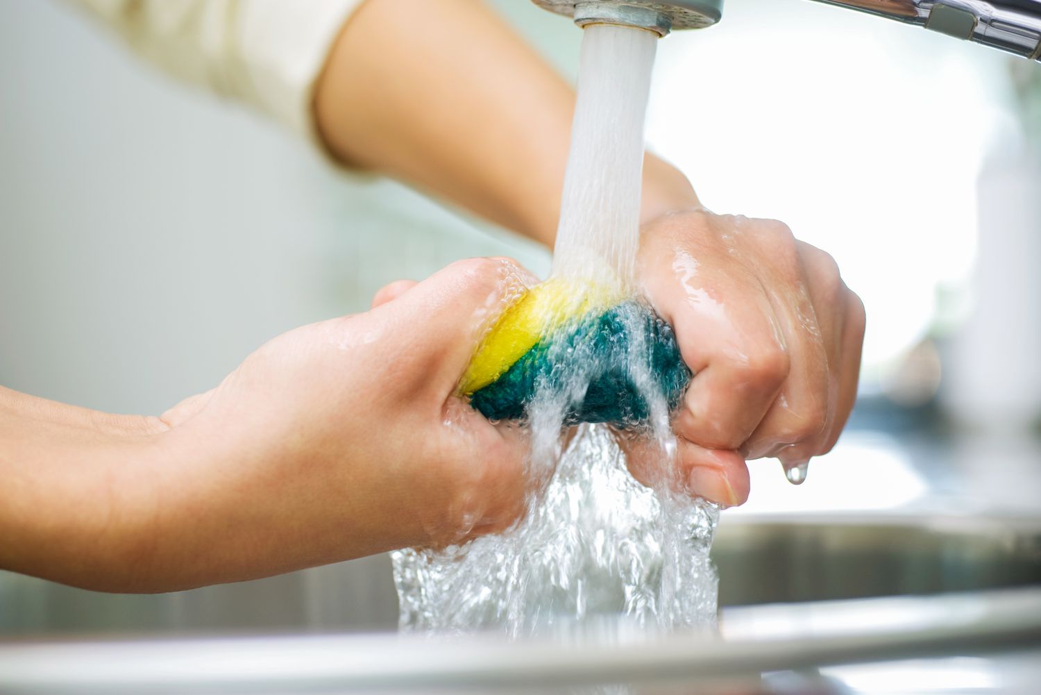 someone wringing out a sponge under running water from a kitchen faucet