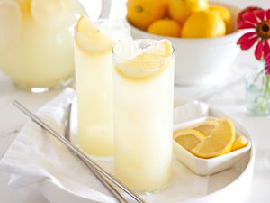 Two glasses of lemonade with lemon slices on a tray, a bowl of lemons, and a vase of flowers in the background