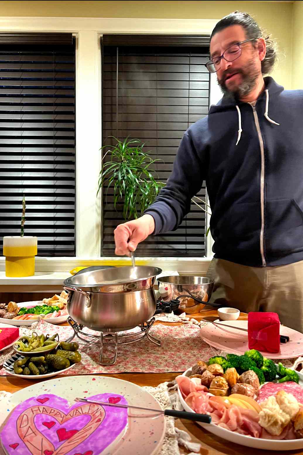 Man stirring the cheese fondue pot on a dining room table set with plates of various dipping foods for a Valentine's Day dinner