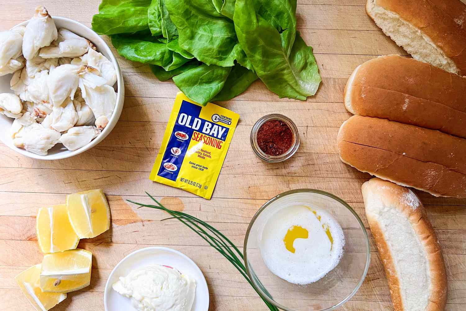 Ingredients for Martha Stewart's crab roll recipe on a wooden surface including crab meat Old Bay seasoning lettuce bread rolls and lemon wedges