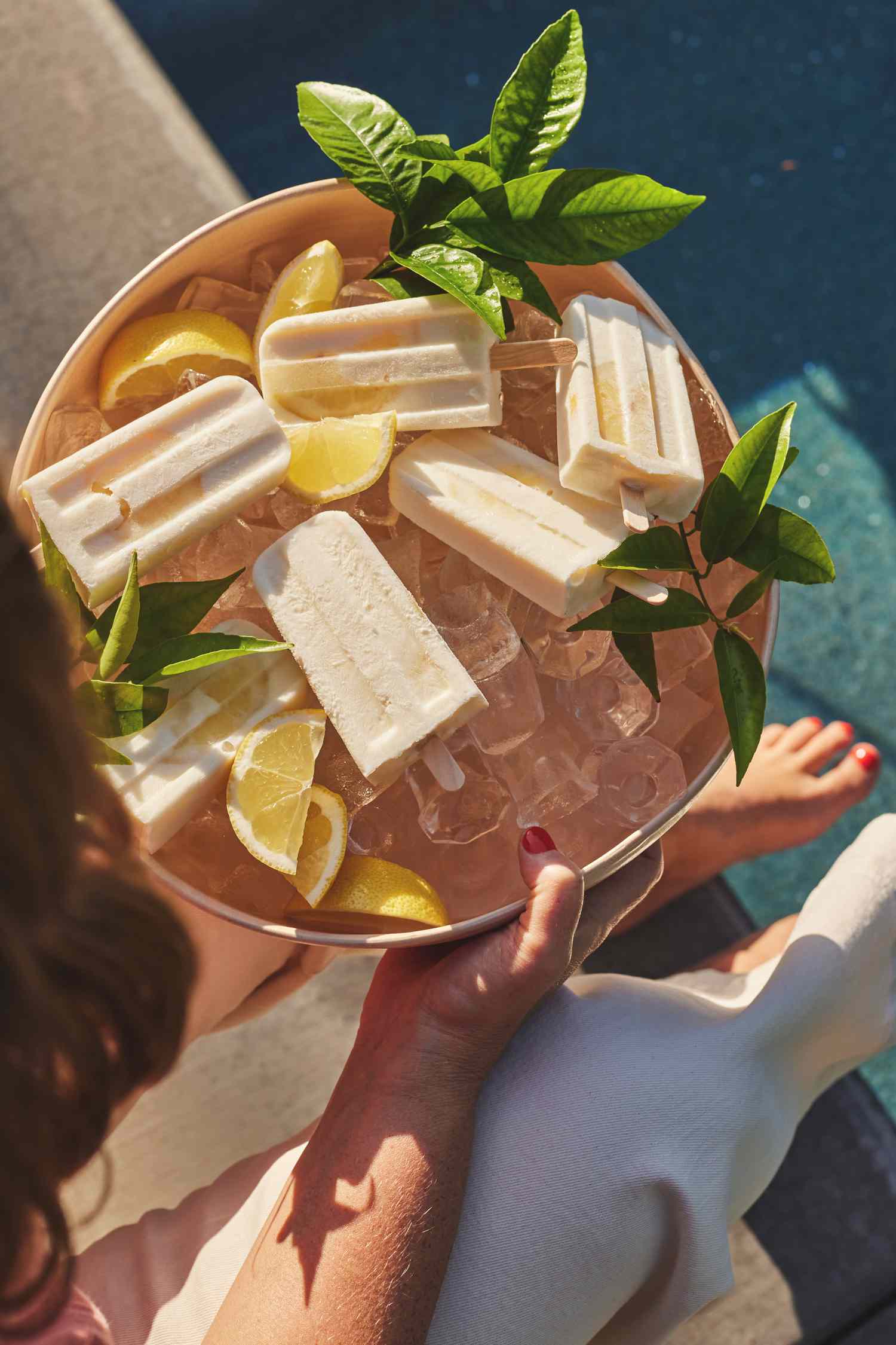 Someone Holding a Bowl of Limoncello Yogurt Pops on Ice Surrounded by Leaves and Lemons Next to a Pool