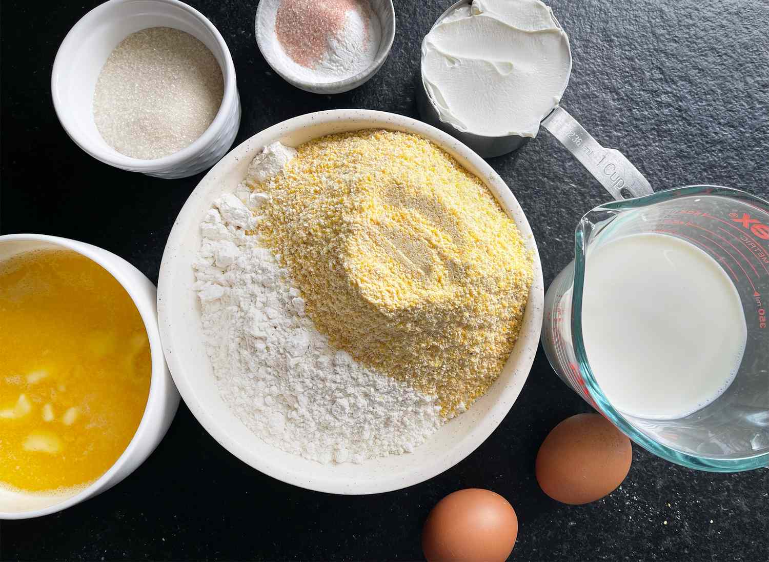 Ingredients for sour cream cornbread including flour cornmeal eggs butter milk sugar baking powder and sour cream on a countertop