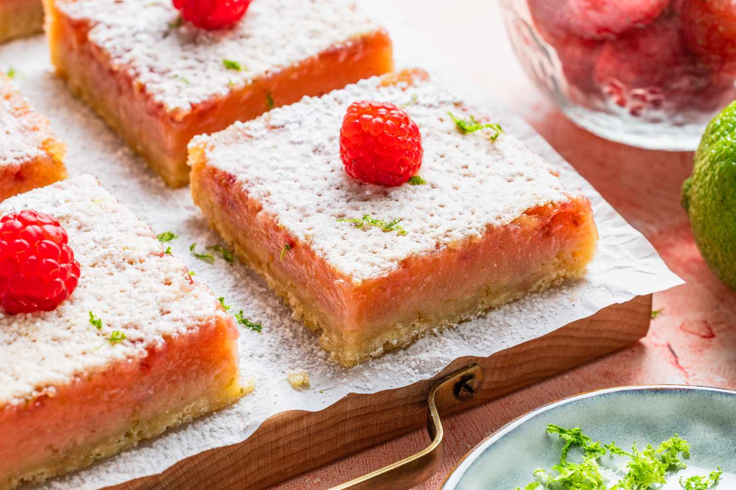 raspberry lime rickey bars on a parchment covered tray at a table setting with a jar of raspberries, a lime, and a small plate with lime zest 