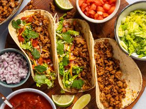 Three tacos with sausage filling on a plate, surrounded by bowls of taco toppings including diced tomatoes, lettuce, onions, and salsa