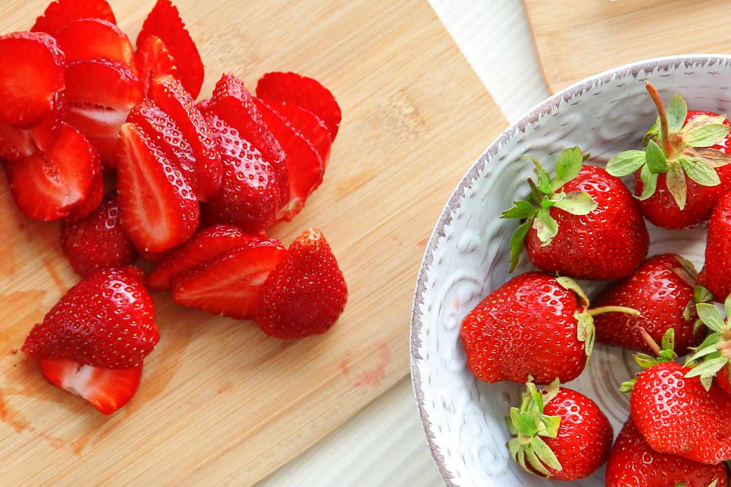 Sliced strawberries on a cutting board beside whole strawberries in a bowl