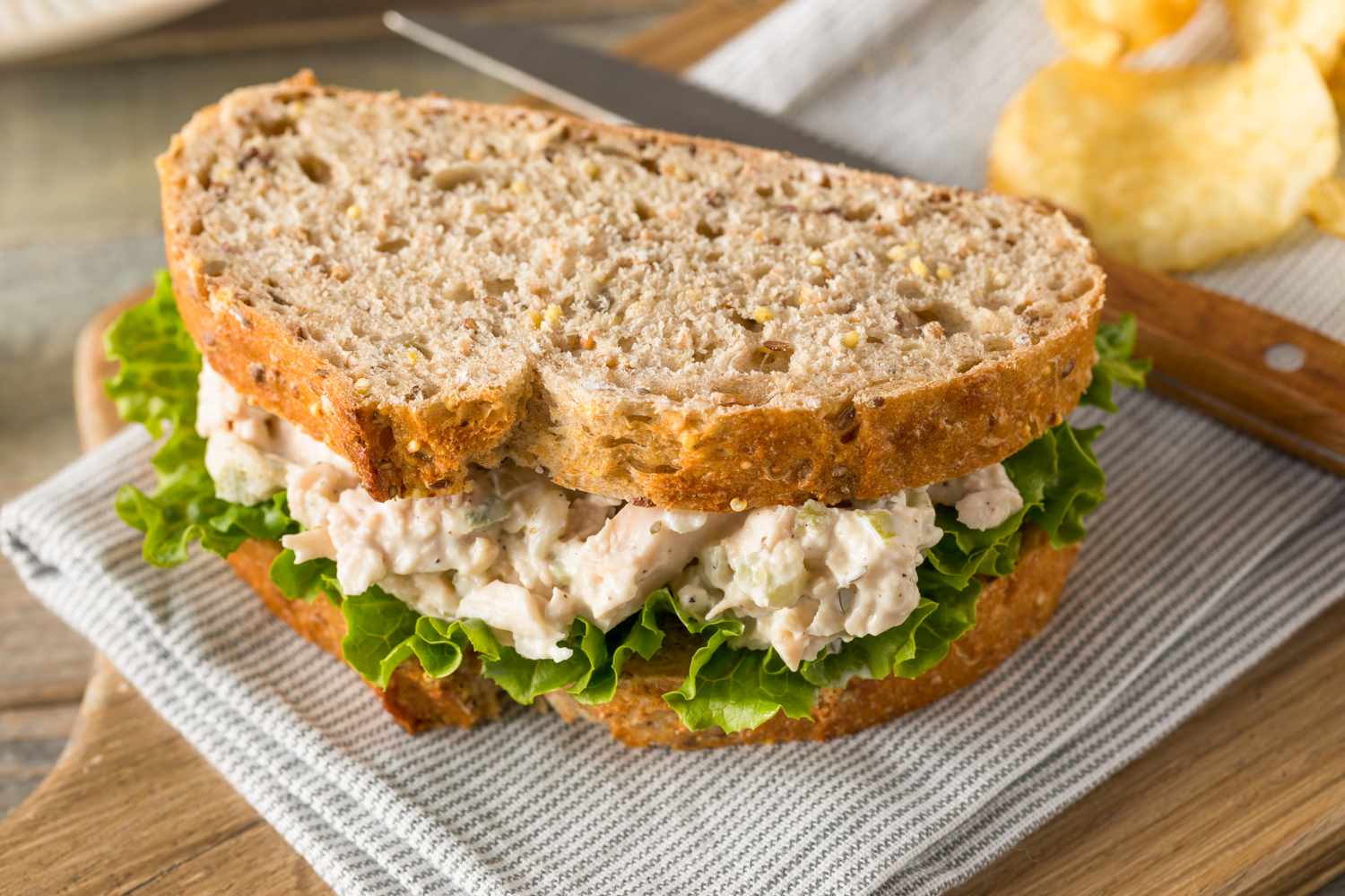 chicken salad sandwich on a table napkin next to a knife and chips, all on a wooden board