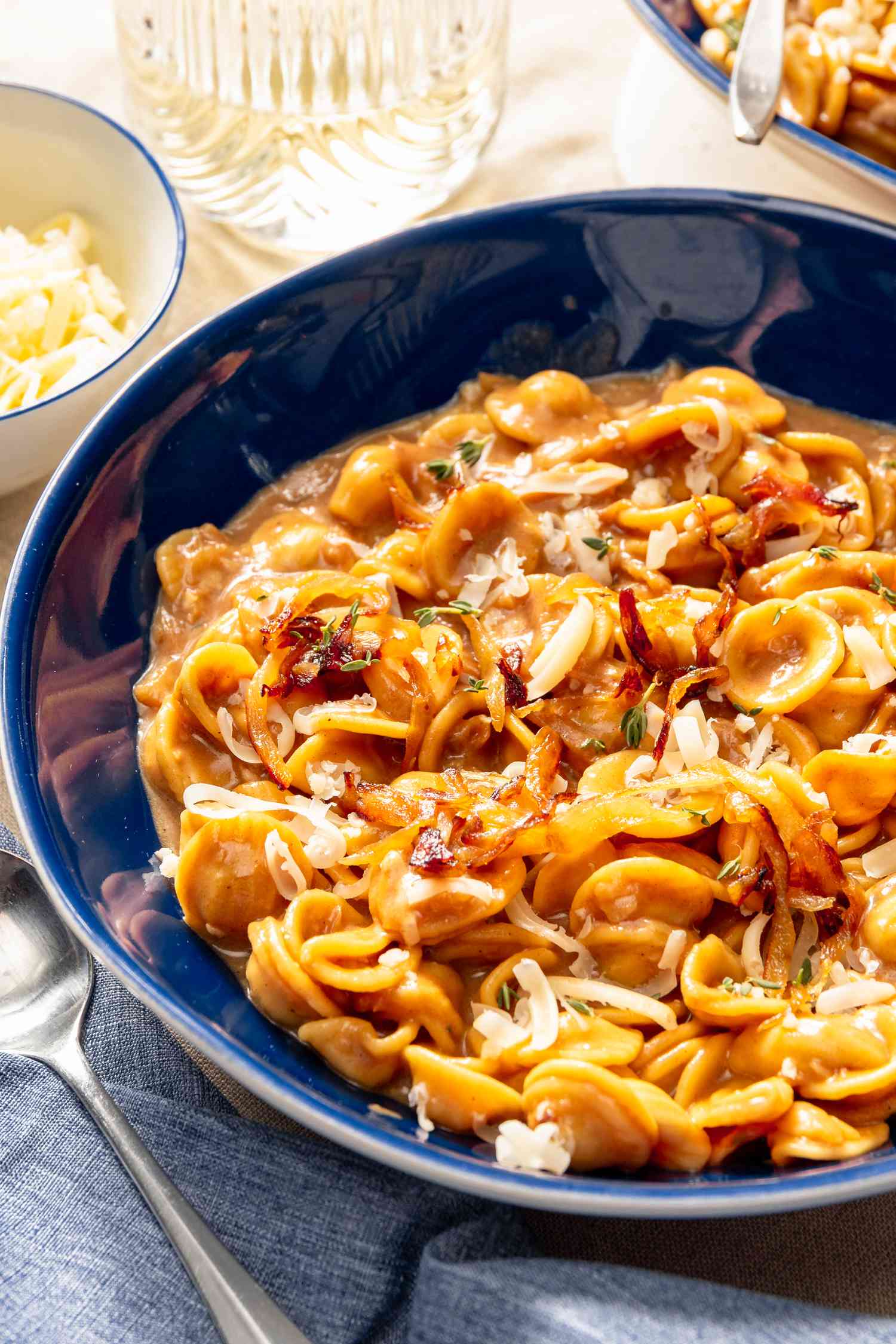 French onion pasta in a bowl at a table setting with another serving in the background, a small bowl of grated cheese, and a glass of water 