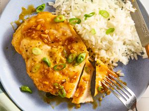 Overhead view of a blue plate with a serving of partially sliced honey soy chicken and white rice topped with green onions along with a fork and knife