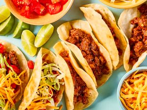 An assortment of tacos with beef shredded lettuce shredded cheese garnishes and lime wedges