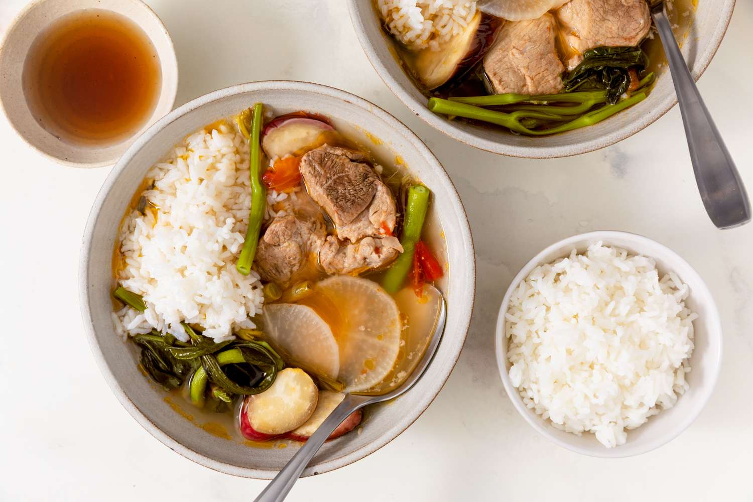 Bowls of pork sinigang with rice and vegetables served on a table alongside a cup of soup