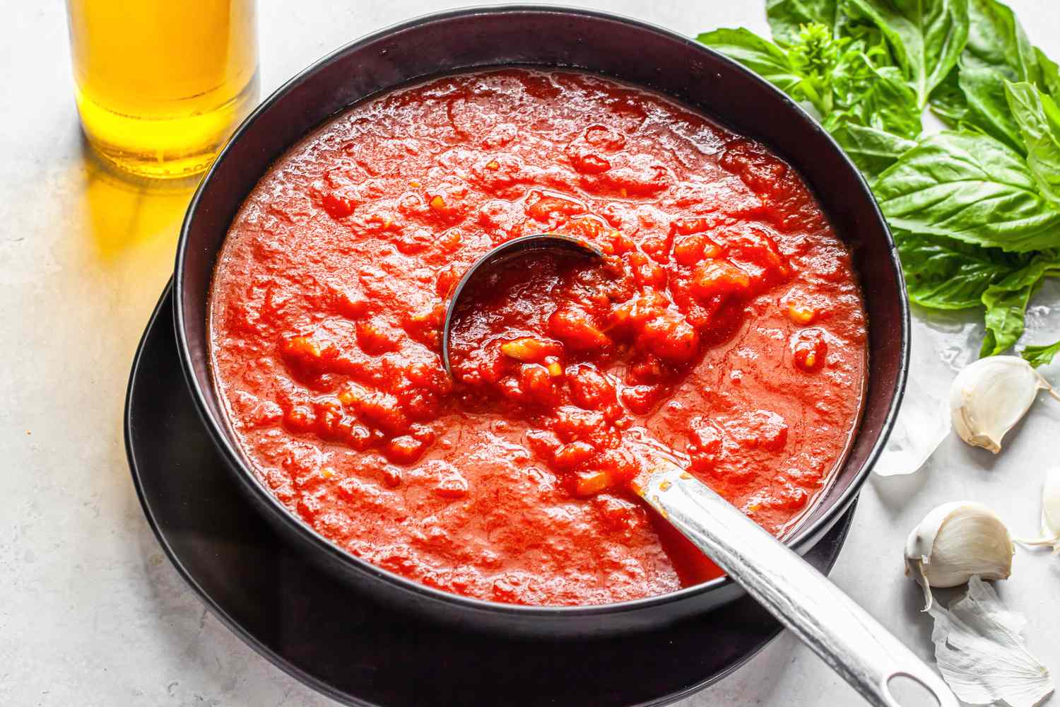 Bowl of Homemade Marinara Sauce With a Ladle, and in the Surroundings, a Bottle of Olive Oil, Garlic Cloves, and Fresh Basil Sprigs