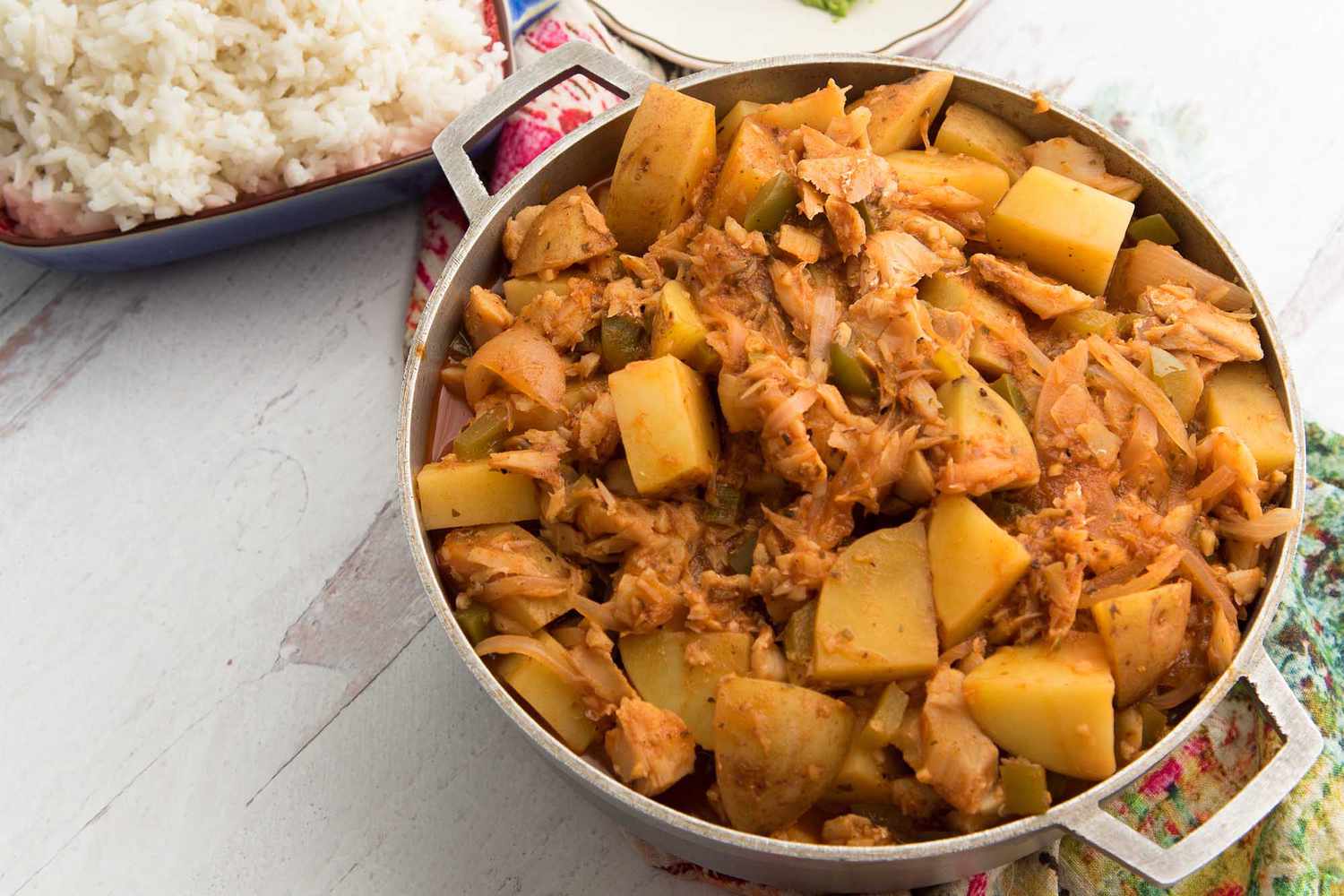 A dutch oven filled with Puerto Rican Stewed Fish is on a white wooden table with a green and pink linen underneath. White rice is in a casserole dish in the upper left corner.