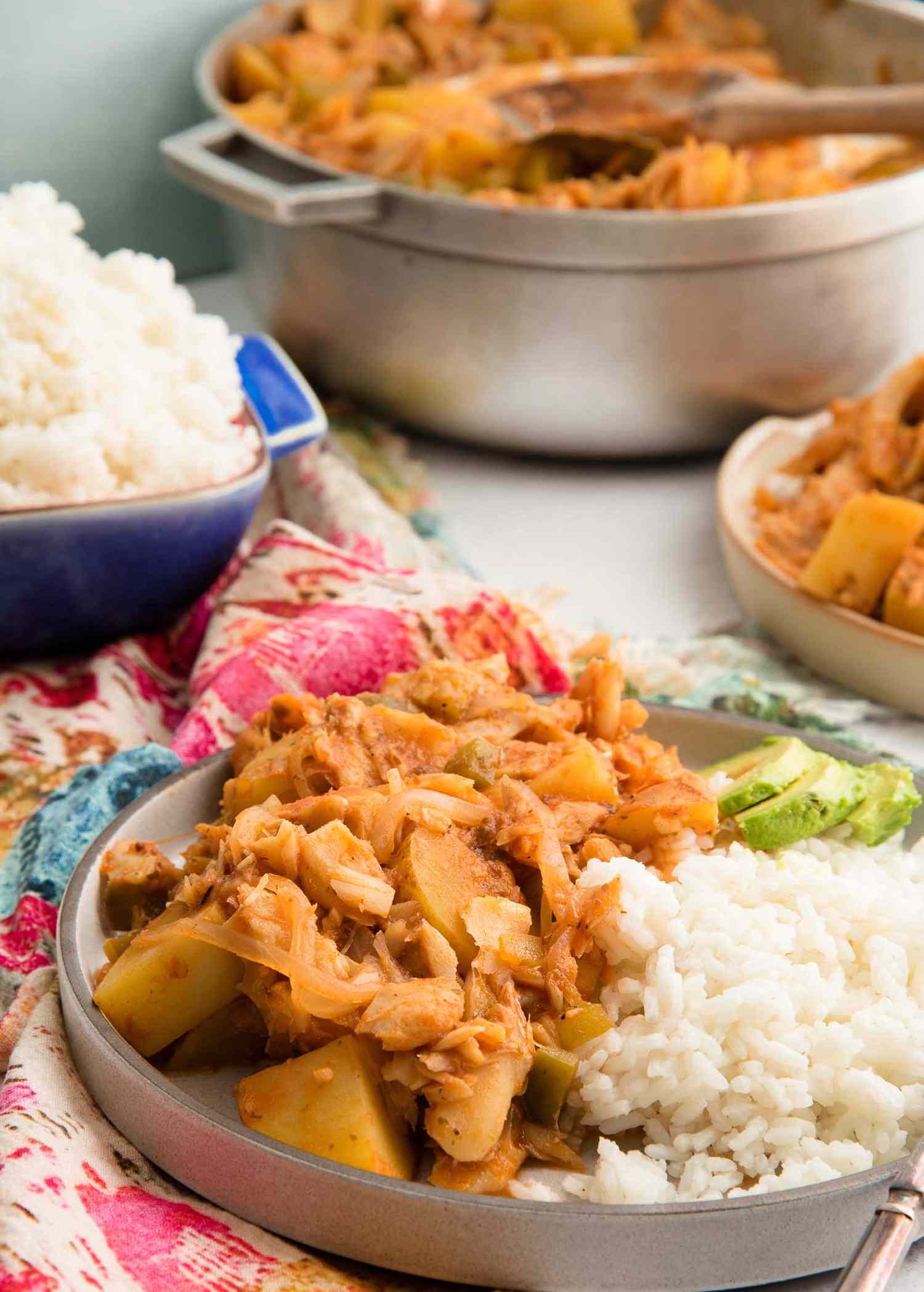 Rimmed grey plate with heaping portion of Puerto Rican Cod Fish. White rice and sliced avocado are on the plate as well. Another plate is to the upper right and a dutch oven filled with the stewed codfish is upper center. A blue casserole dish is heaped with white rice and is above the plate to the left. A pink and blue patterened linen is under the plate and rice dish.