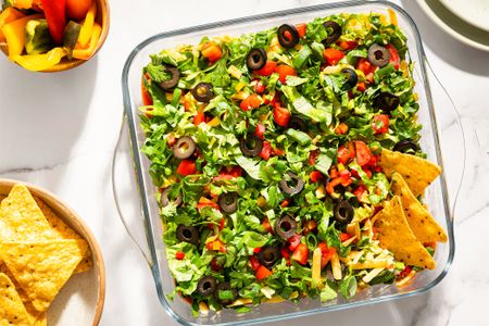 Layered nacho dip in a glass dish with toppings like lettuce tomatoes and olives served with tortilla chips
