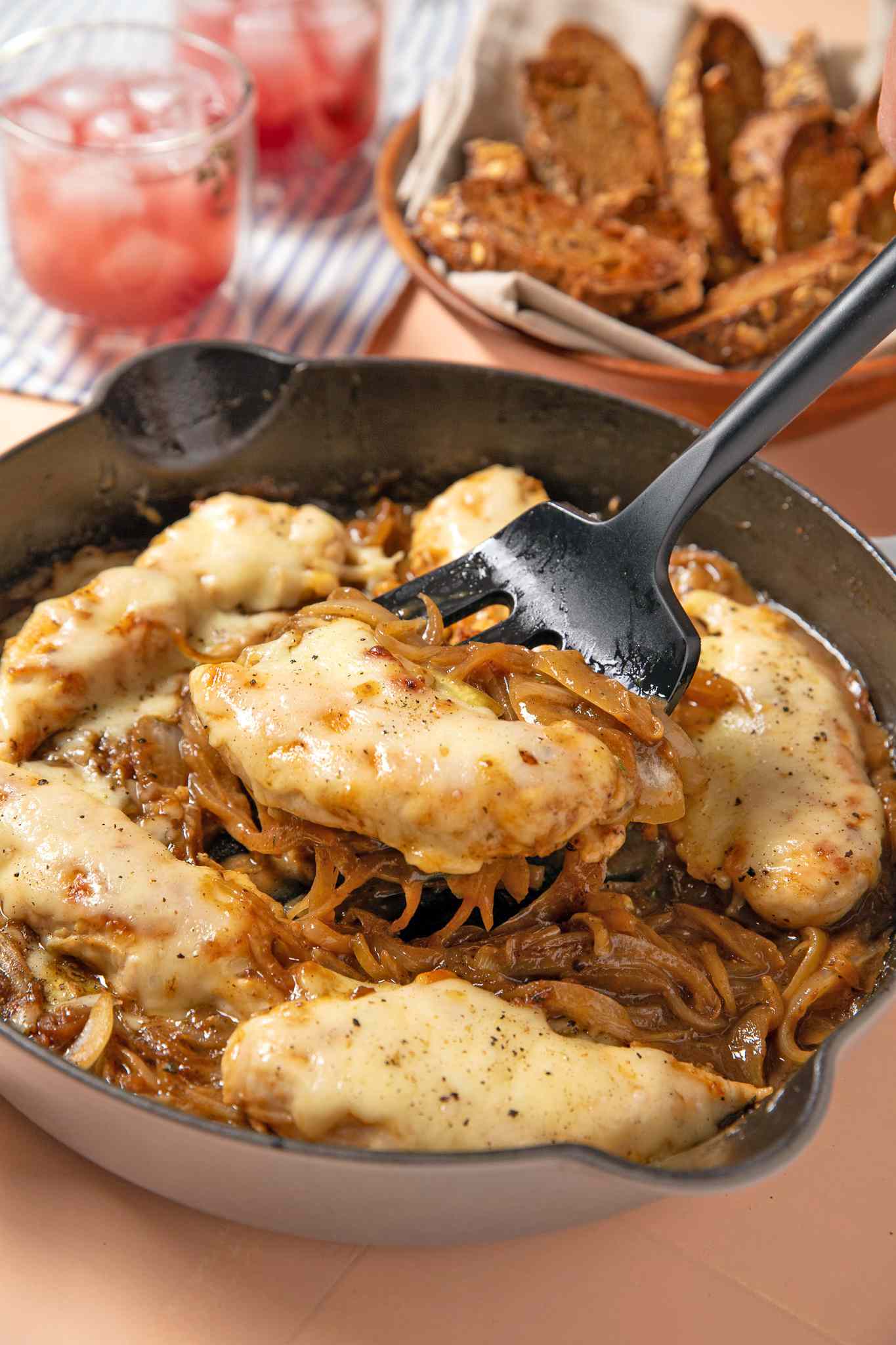 french onion chicken lifted from a skillet using a spatula, and in the background, glasses and a bowl of bread