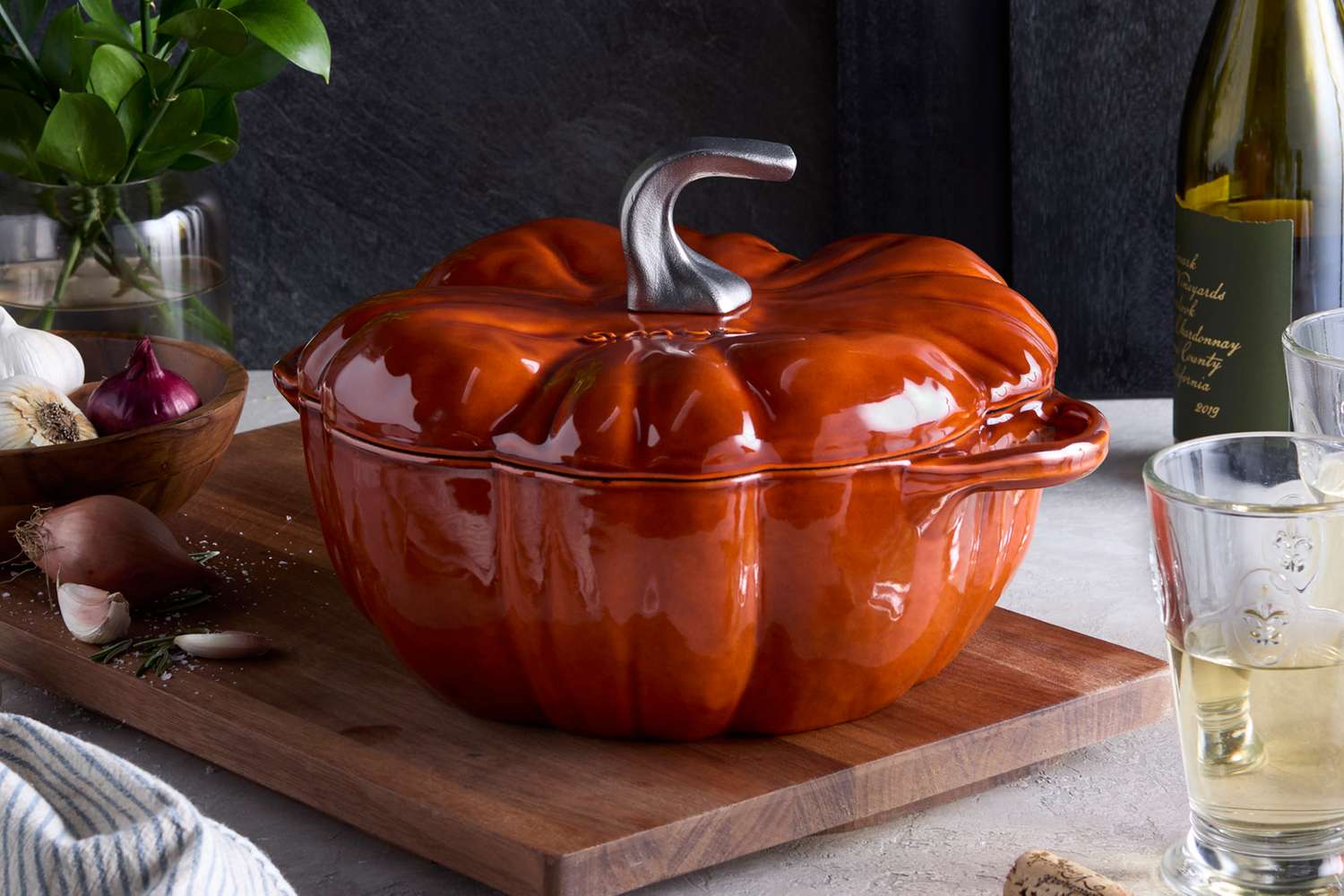 A cast iron pot shaped like a pumpkin on a wooden board surrounded by wine and cooking ingredients