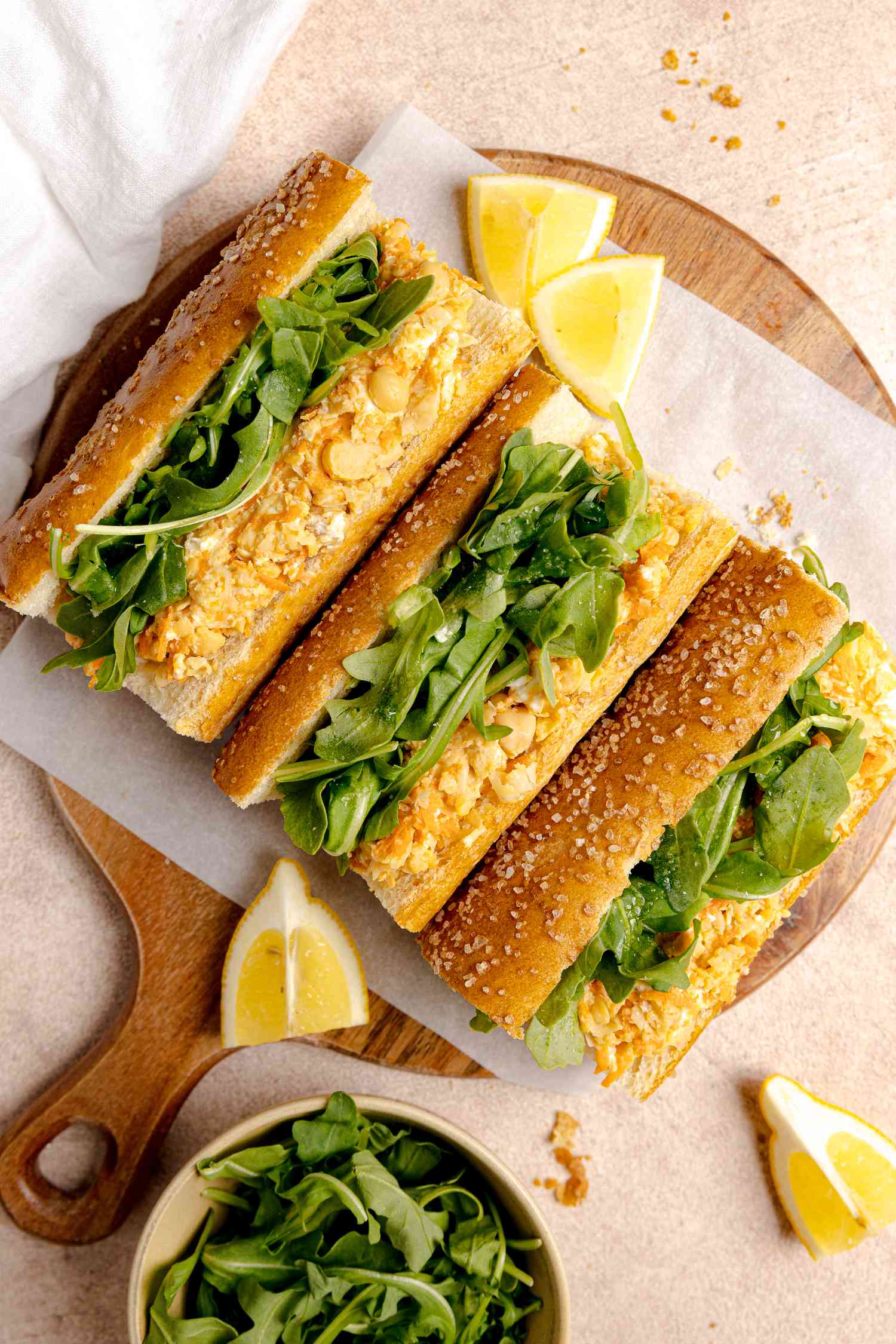 Chickpea Paninis on Parchment Paper over a Circular Wooden Board Surrounded by Lemon Wedges and a Bowl of Arugula