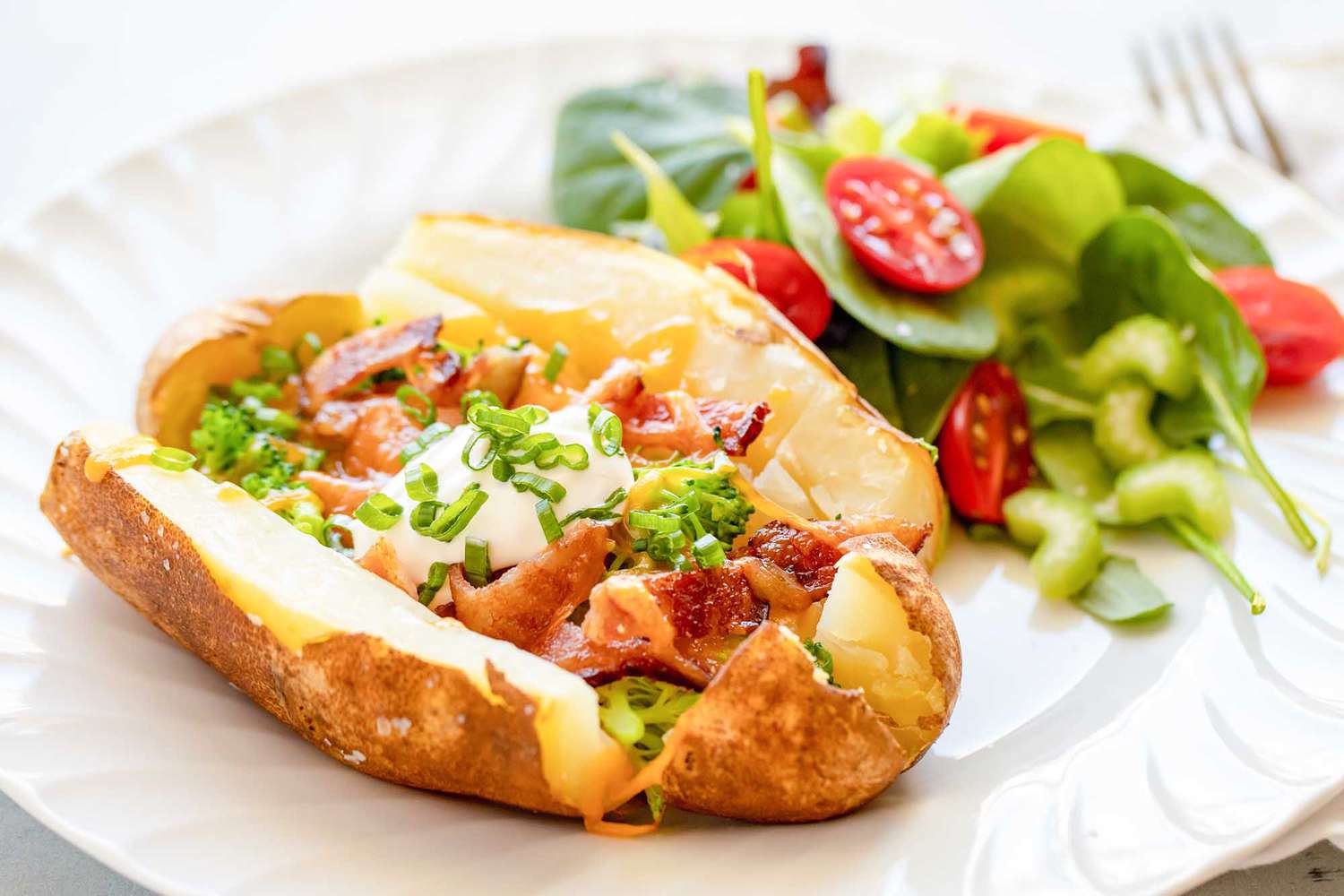 Cheesy, broccoli, bacon loaded baked potato on a white plate with a salad of spinach and tomato behind it on a white plate. The potato is sliced down each side and the top and bottom. Chopped bacon, broccoli, scallions and shredded cheddar cheese are on the potato. A spoonful of sour cream is in the middle of the potato. A fork is to the right of the plate.