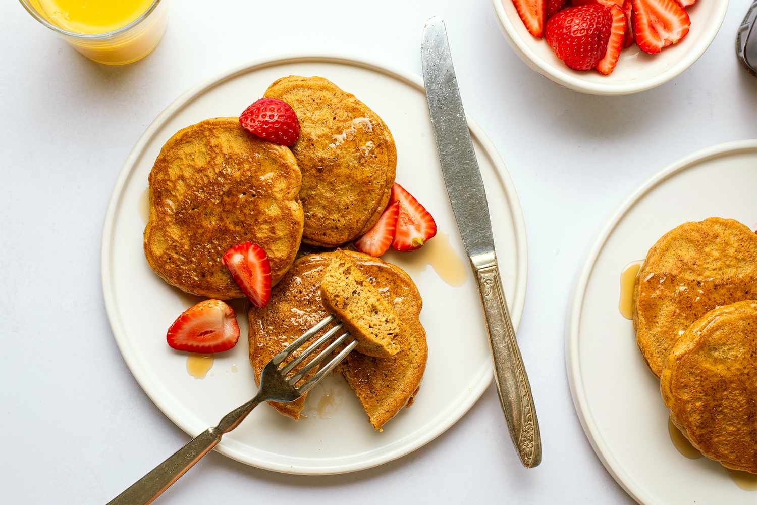 A plate of almond butter pancakes garnished with strawberries a fork with a slice of pancake and a nearby bowl of strawberries