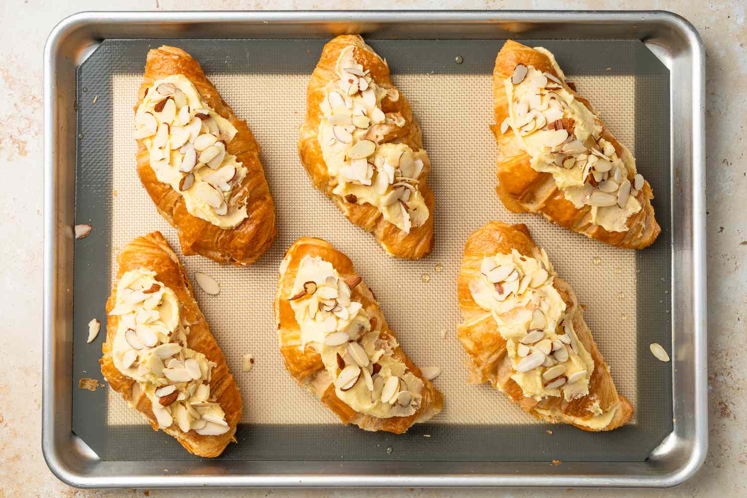Almond Croissants Assembled on a Silicone Mat Lined Baking Sheet and the Tops of Each Croissant Is Covered in Frangipane and Almond Slices