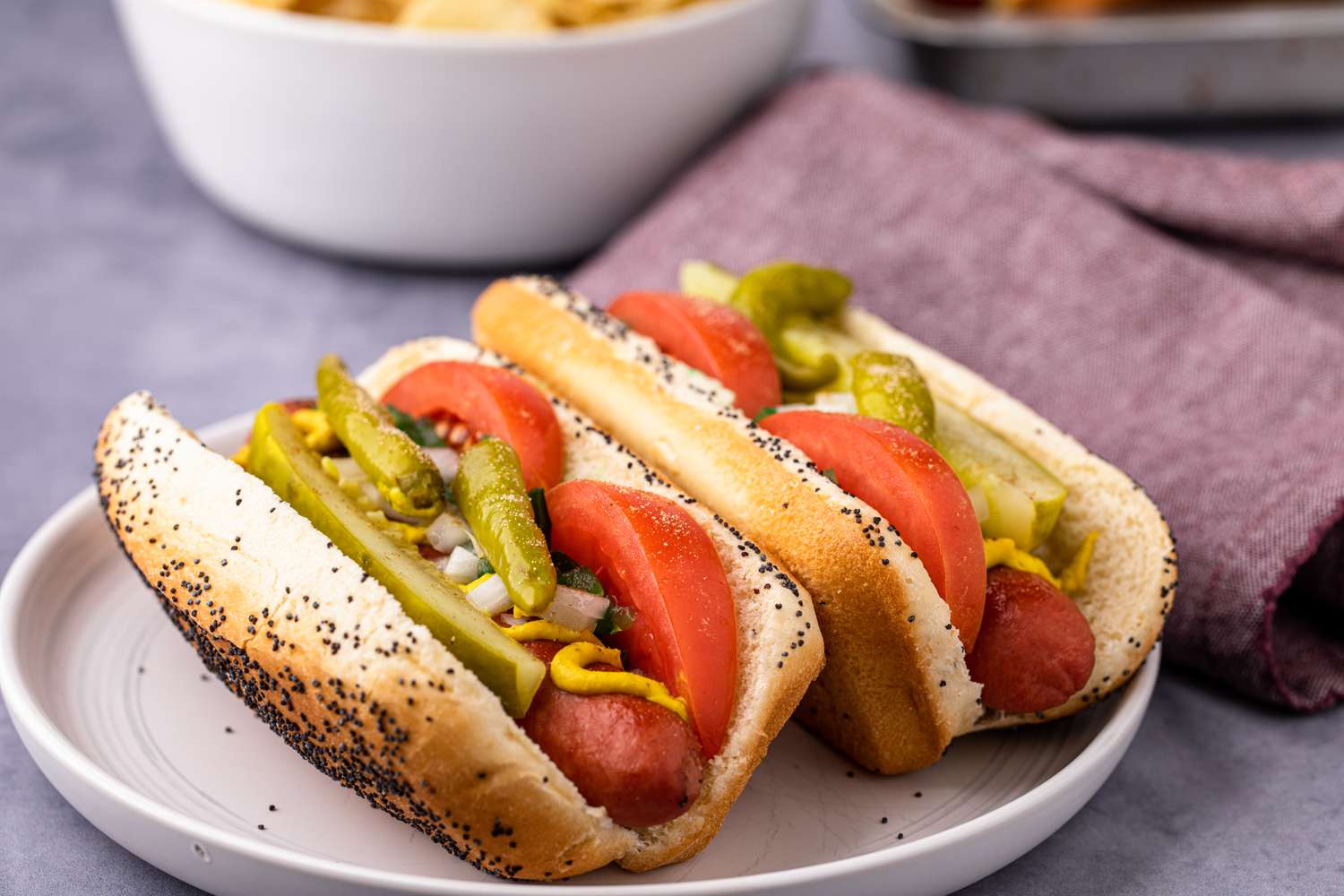 All-beef hot dog with toppings on a plate with a bowl of chips behind it.