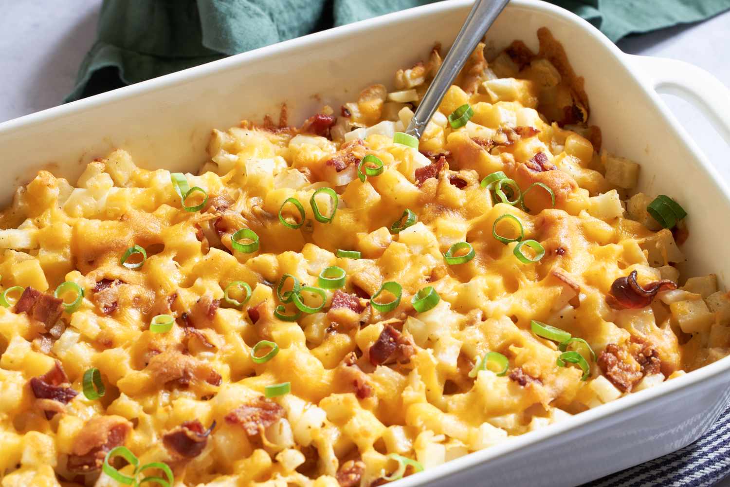 Baked dish of diced potatoes topped with melted cheese bacon bits and chopped green onions served in a rectangular casserole dish