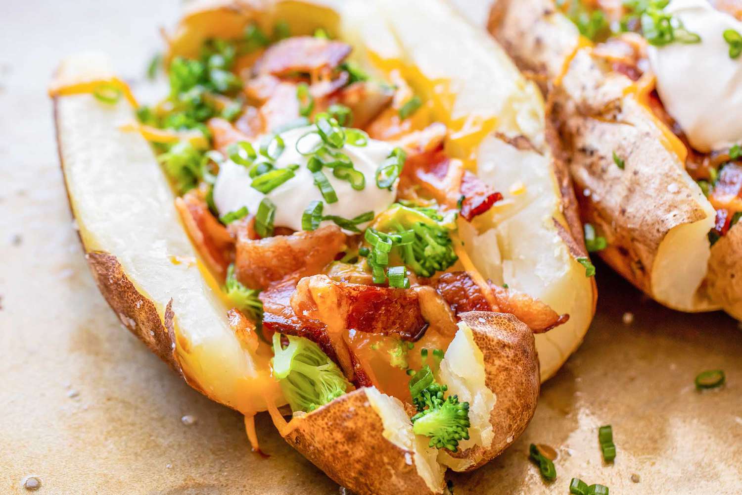 Side view of quick and easy loaded baked potatoes on a worn baking sheet. Along with cheddar cheese, broccoli and bacon, the potato has a spoonful of sour cream and chopped scallions. A second potato is partially visible to the right.